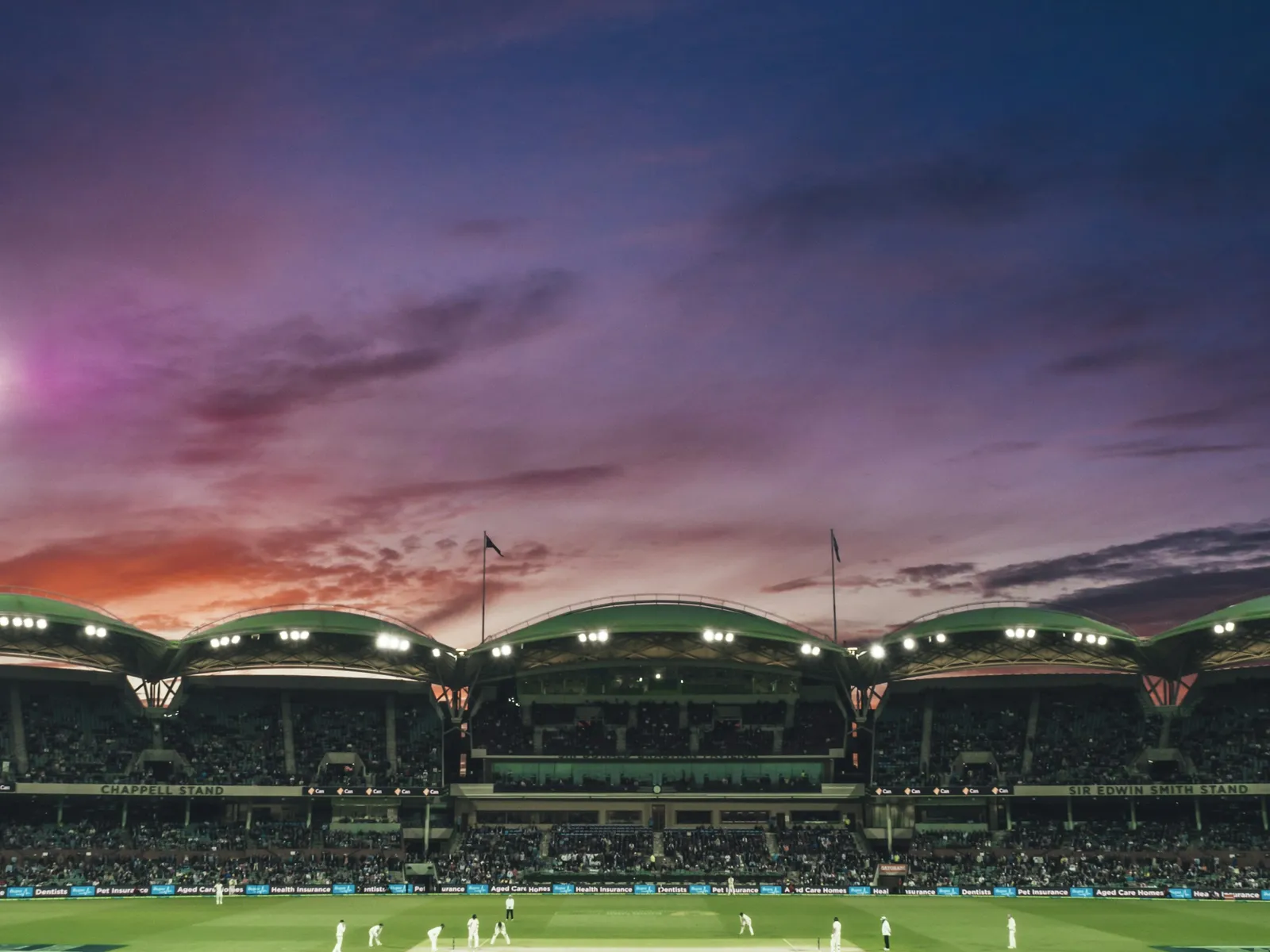 A floodlit cricket ground at dusk, seen from the stands